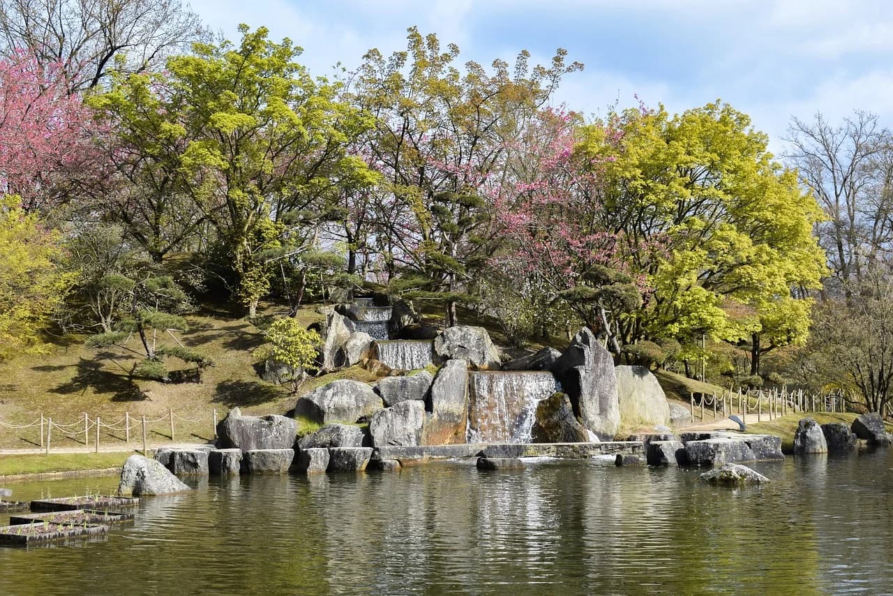 Water cascades over large rocks in the Japanese Garden of Hasselt, surrounded by a variety of trees with colorful spring blossoms.