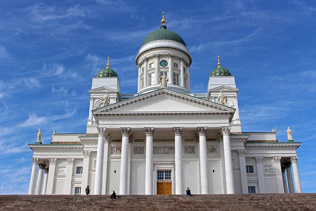 The iconic Helsinki Cathedral is viewed from below, its impressive white columns and green domes rising majestically against a bright blue sky.