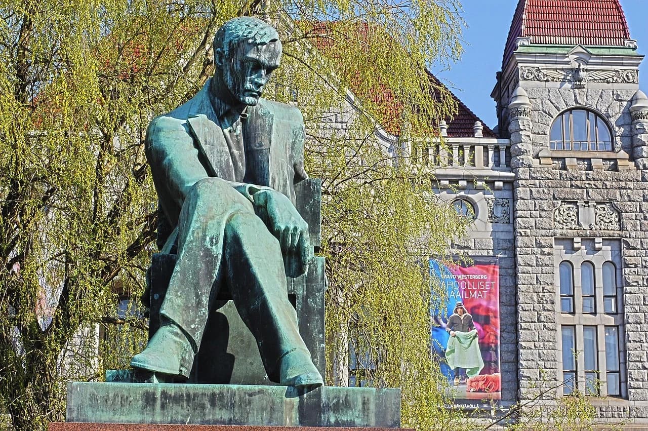 A bronze statue of Finnish composer Jean Sibelius sits thoughtfully in a park, with a historic stone building in the background.