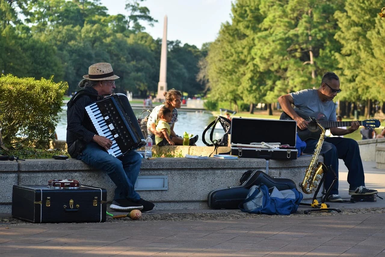 Two street musicians, one playing an accordion and the other a guitar and saxophone, perform by a pond in a park.