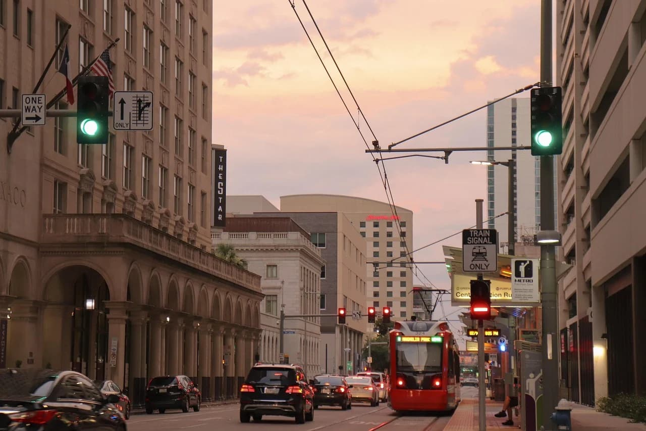 A tram travels along a city street at sunset, with tall buildings and traffic creating a modern, urban scene.