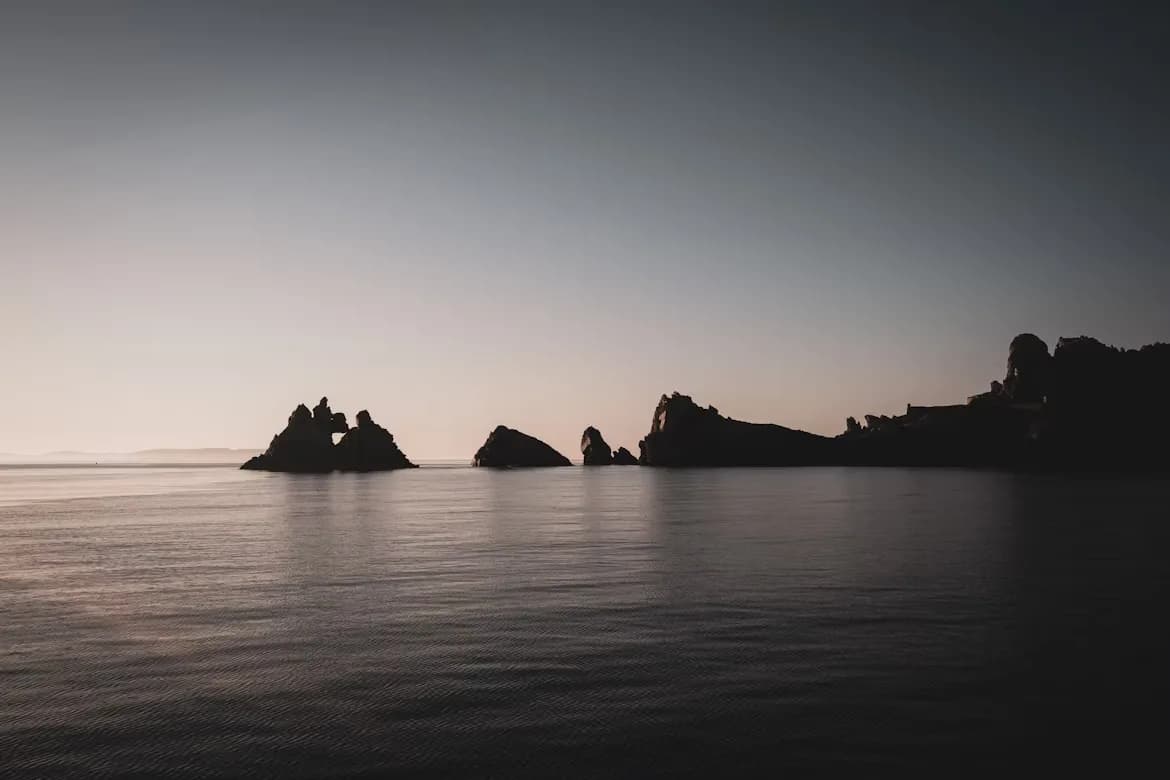 A serene, moody image of jagged rock formations in the sea, silhouetted against a setting sun.