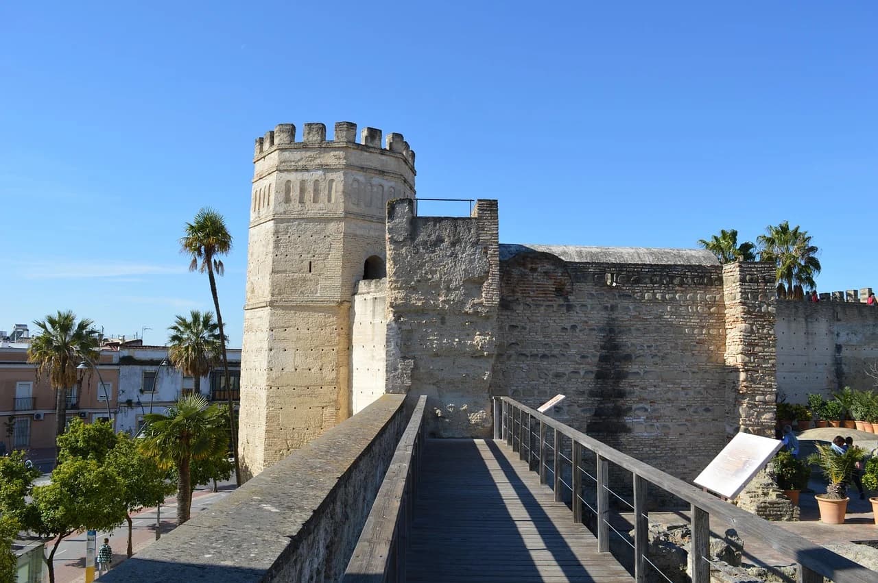 The Alcazar of Jerez de la Frontera, a historic Moorish fortress, features a prominent tower and fortified walls against a bright blue sky.