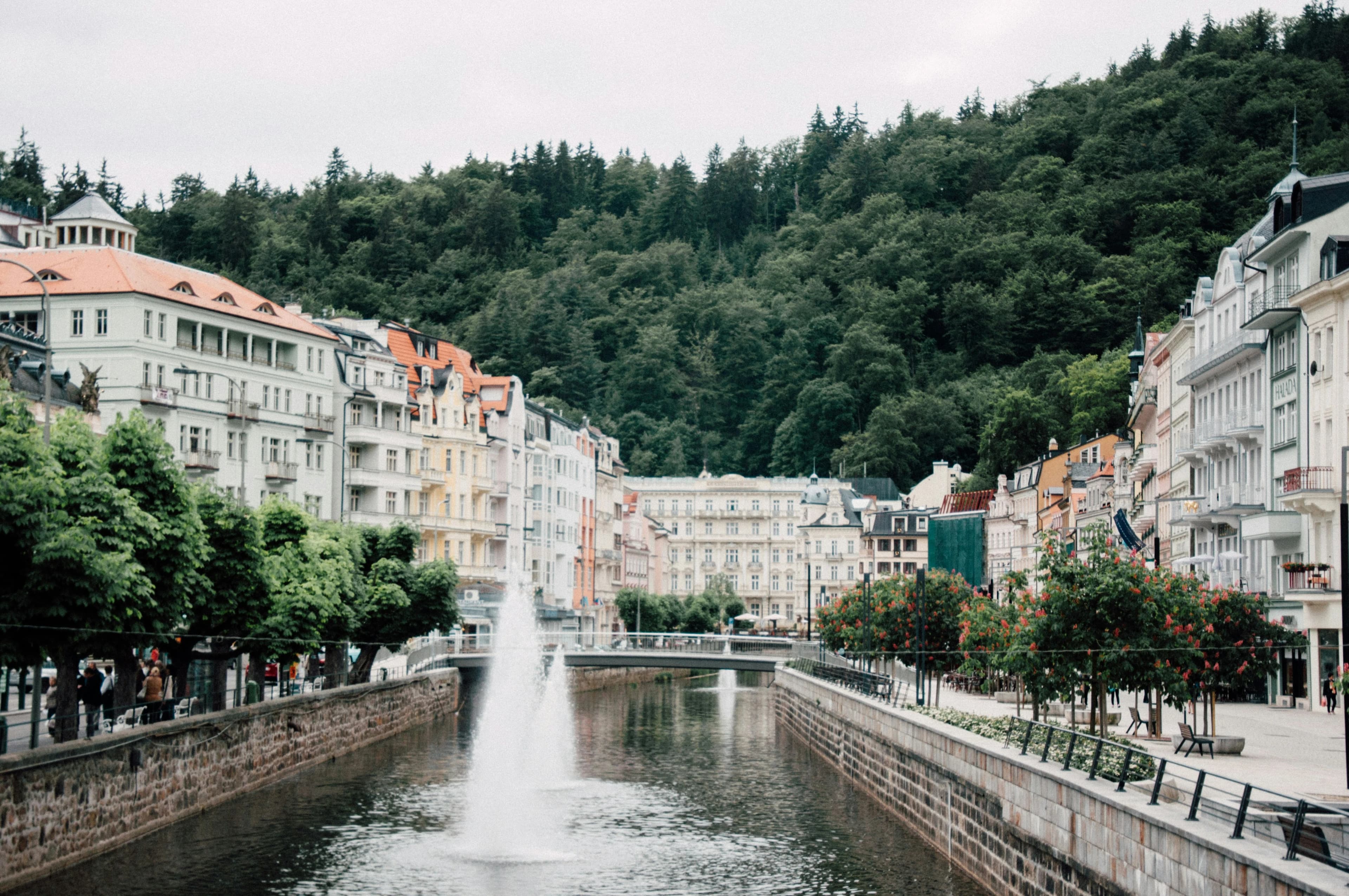 A beautiful fountain sprays water into the river that runs through the heart of Karlovy Vary, lined with trees and historic buildings.