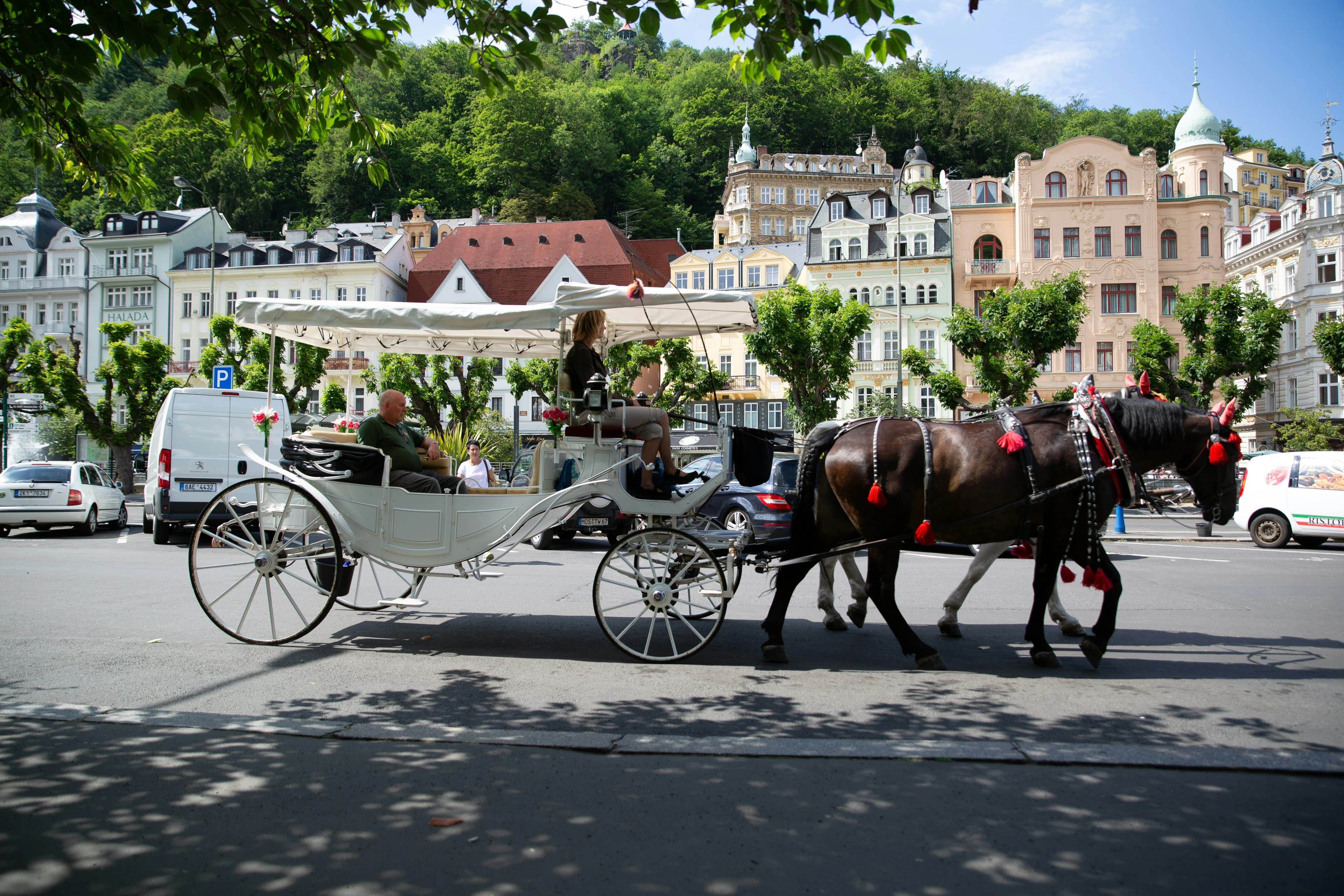 A horse-drawn carriage passes along a busy street, with the elegant historic buildings and lush green hills of Karlovy Vary in the background.