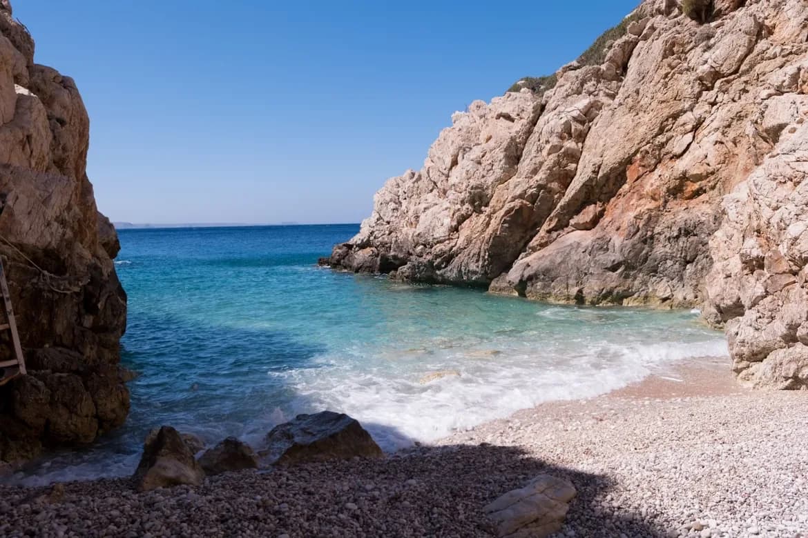 A secluded pebble beach with crystal-clear turquoise water is surrounded by dramatic cliffs.