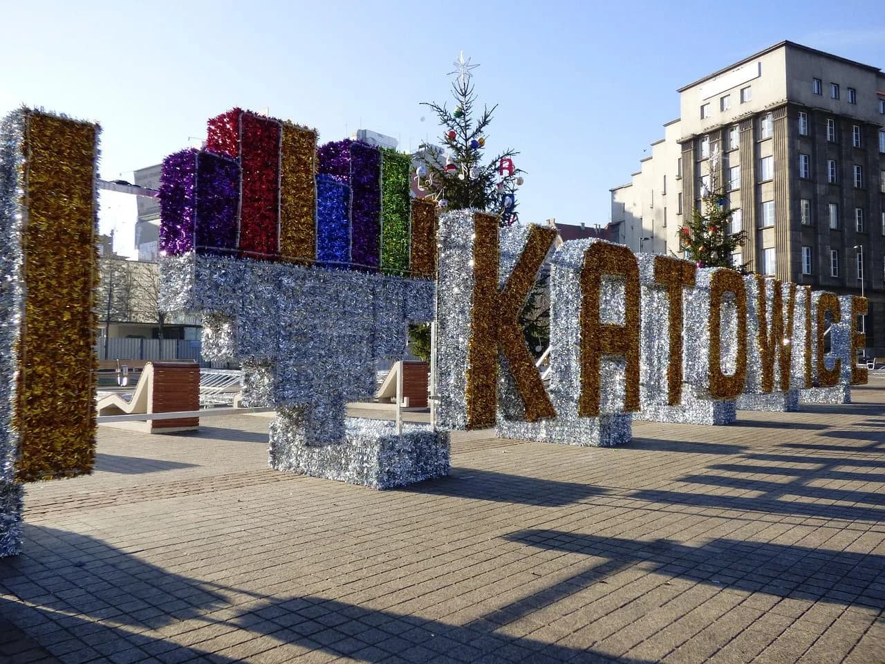 A large, tinsel-covered sign spells out "Katowice" in a public square, with decorated Christmas trees in the background.
