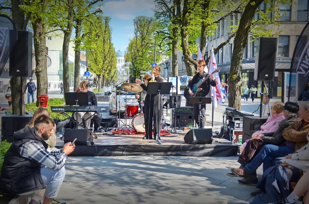A live band performs on an outdoor stage in a public square, entertaining people relaxing on benches under the trees.