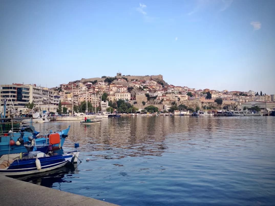 The bustling harbor of Kavala is filled with fishing boats, with the historic city and a castle on the hill in the background.