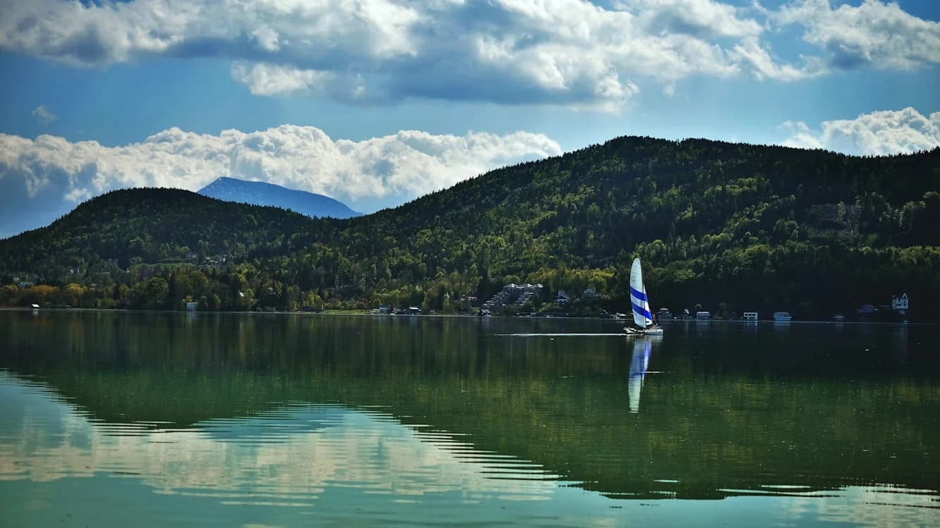 A sailboat with a striped sail glides across the calm waters of a lake, with green, forested mountains in the background.