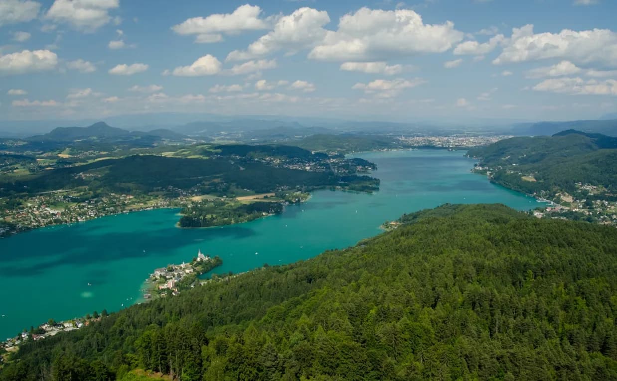 An aerial view captures the turquoise waters of Lake Wörthersee, with a small town and a church steeple on a peninsula.