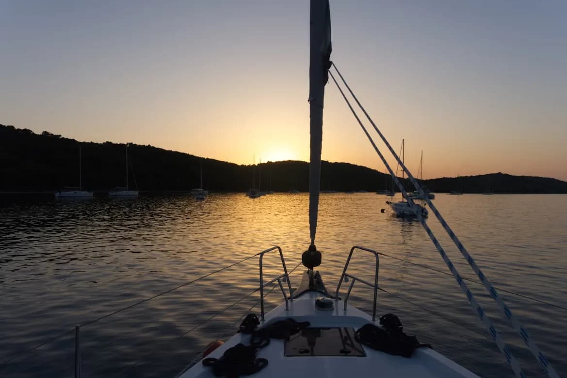 A serene sunset is viewed from the deck of a sailboat, with other boats anchored in a calm bay of the Kornati Islands.