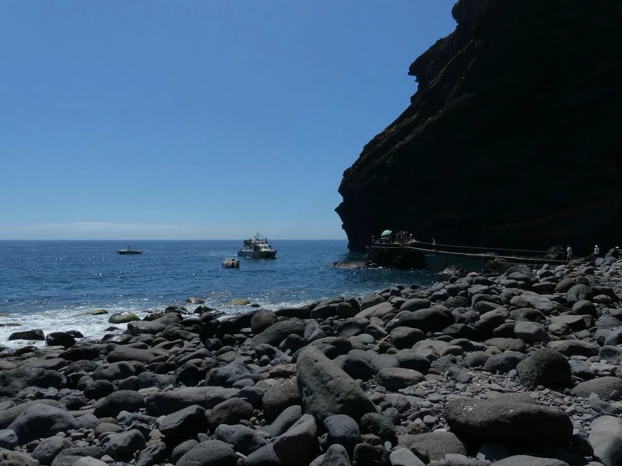 A boat is moored on a rocky beach, with a long wooden walkway and a towering cliff in the background.