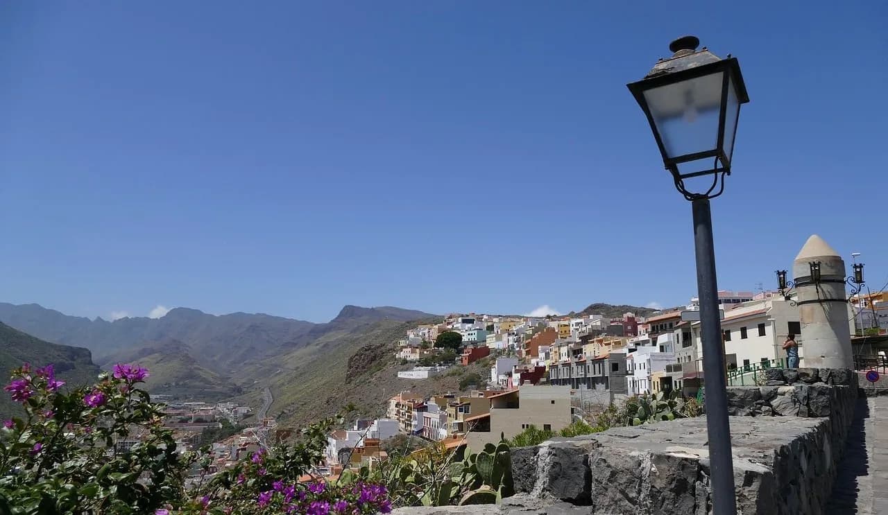 The colorful houses of a town on La Gomera are nestled in a green valley with a lamppost and blooming flowers in the foreground.