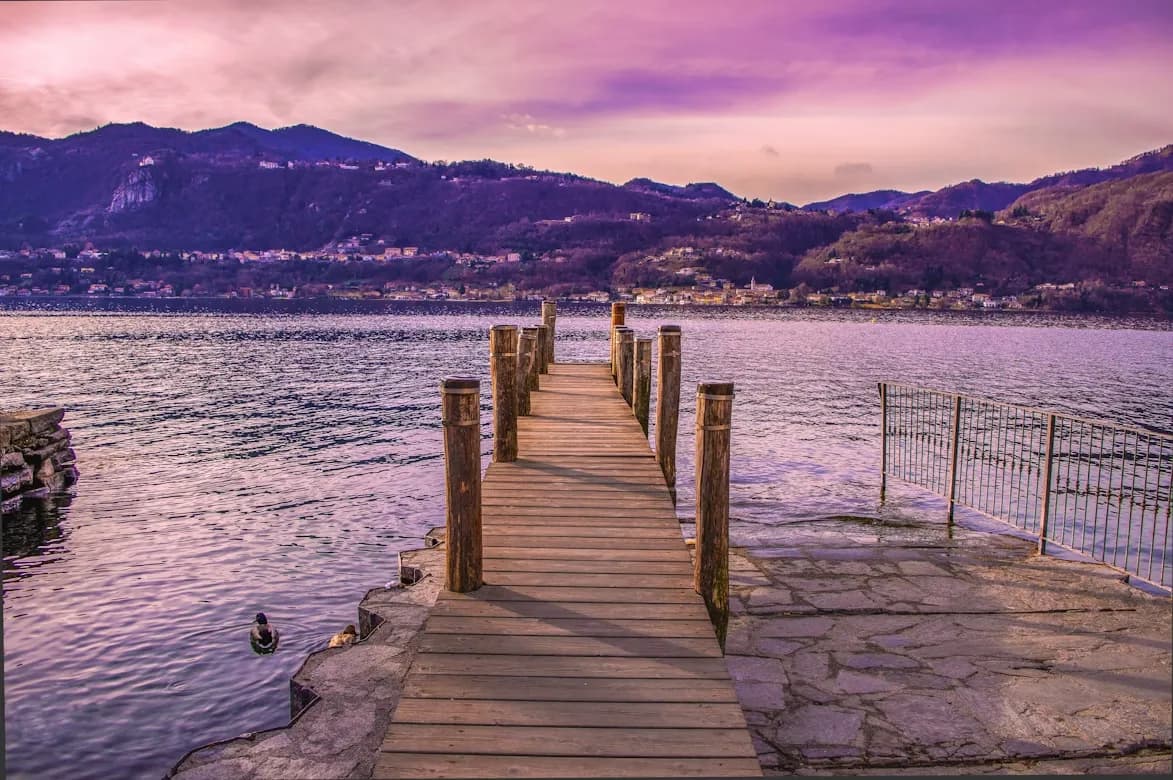 A wooden pier stretches out into the calm, purple-hued waters of Lake Orta, leading toward the mountains in the distance.