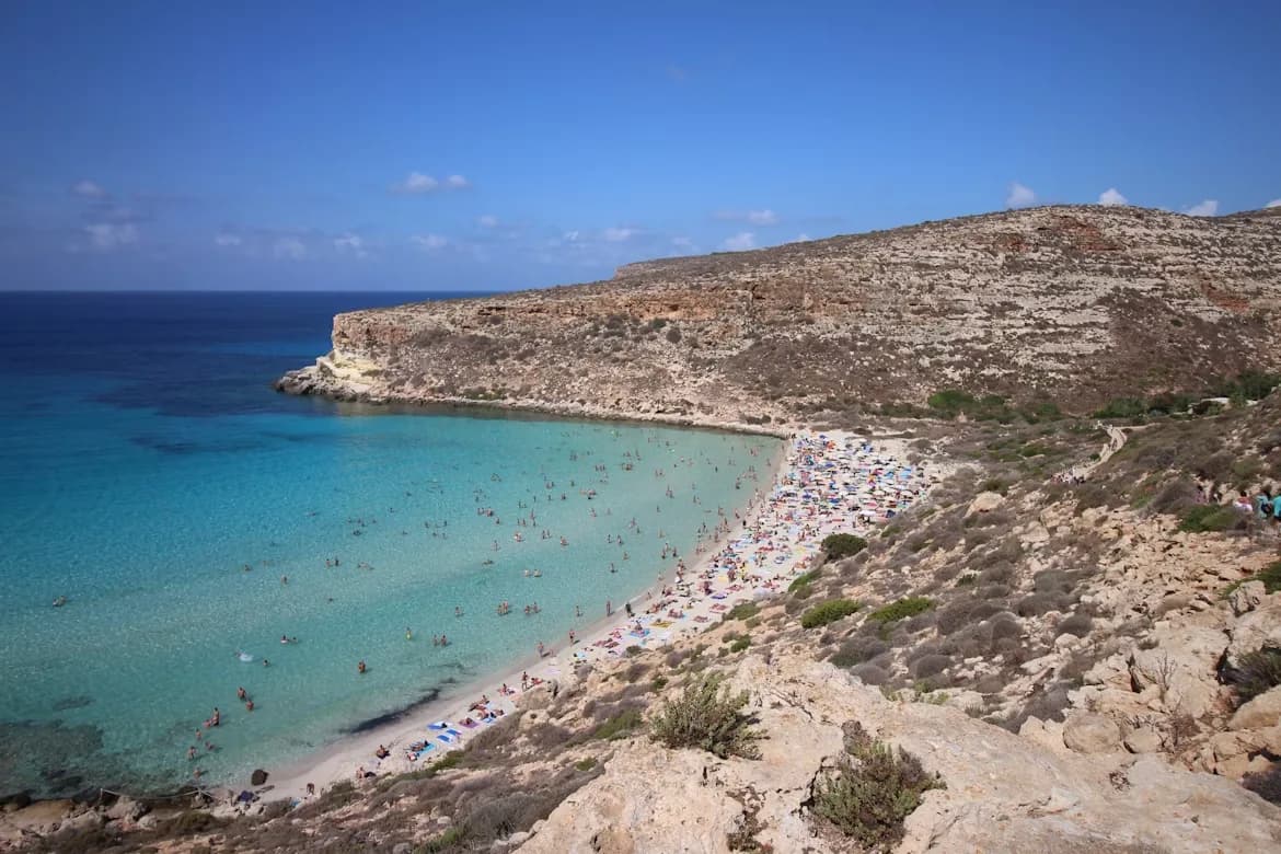A bustling beach with people swimming and sunbathing is nestled in a cove with crystal-clear turquoise water and rocky cliffs.
