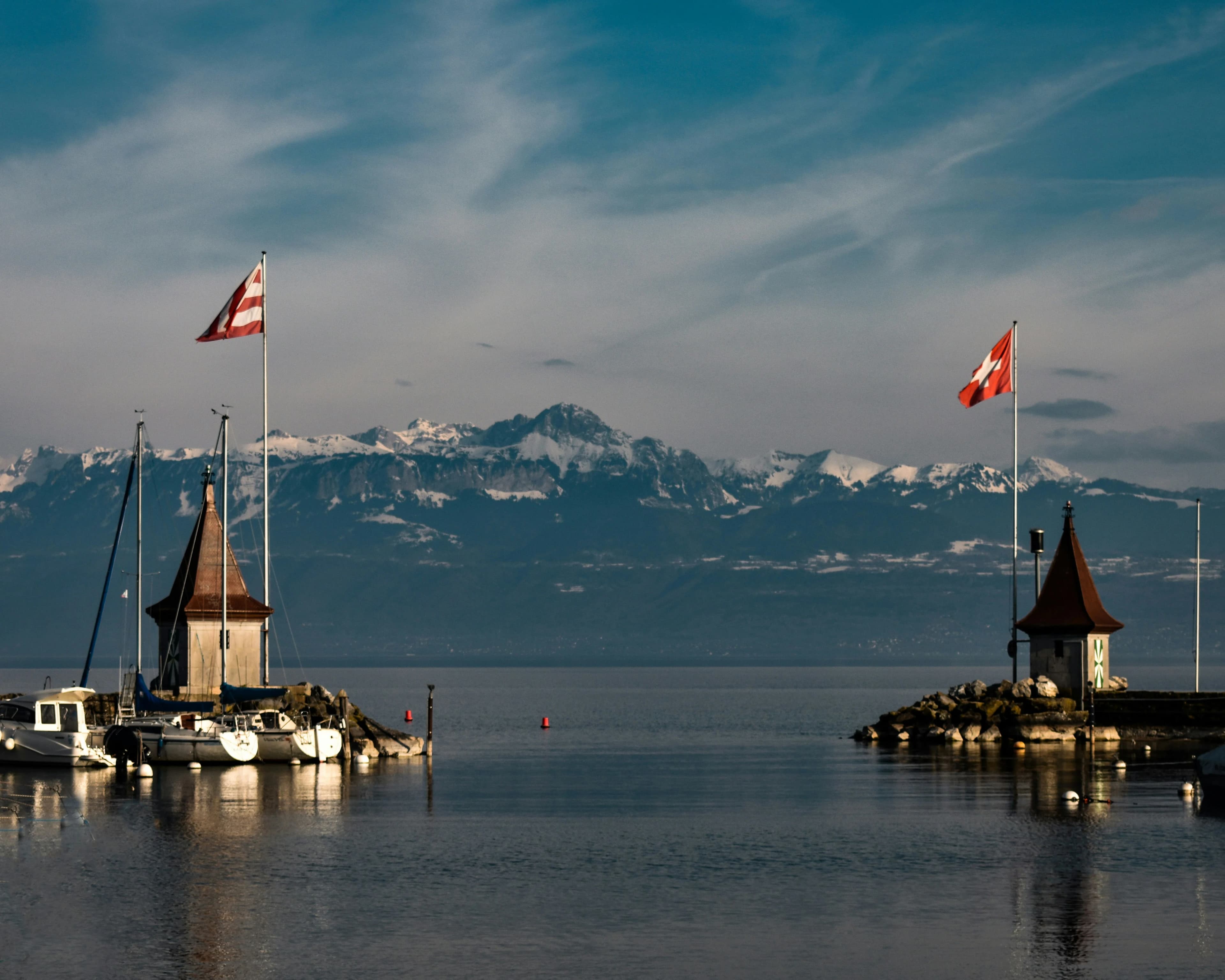 Small harbor houses and sailboats frame a stunning view of the snow-capped Swiss Alps across the peaceful waters of Lake Geneva.