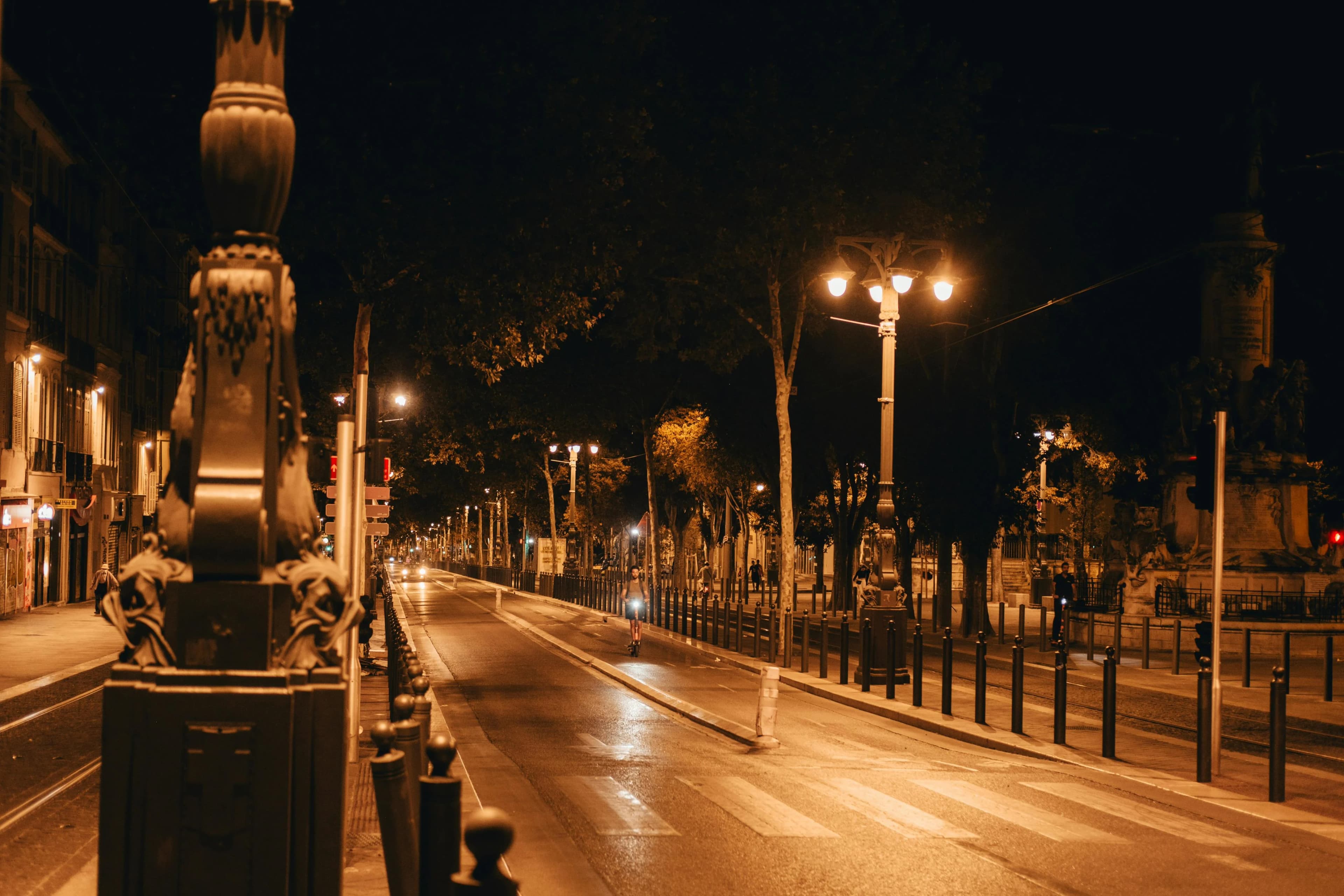 A cyclist rides down a quiet, illuminated street at night, with streetlights casting a warm glow on the buildings.