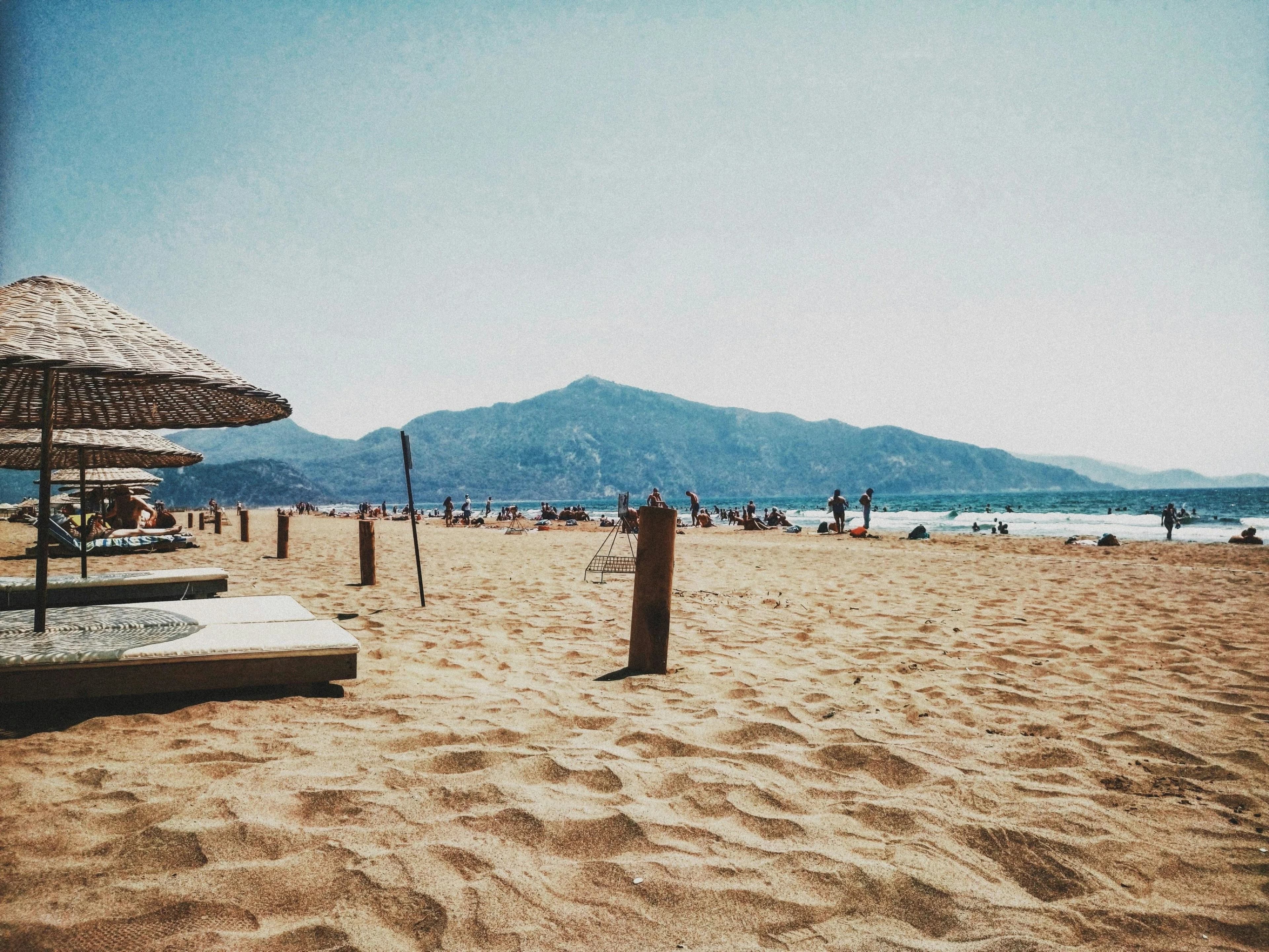 People relax on a sandy beach under thatched umbrellas, with a large, forested mountain looming in the background.