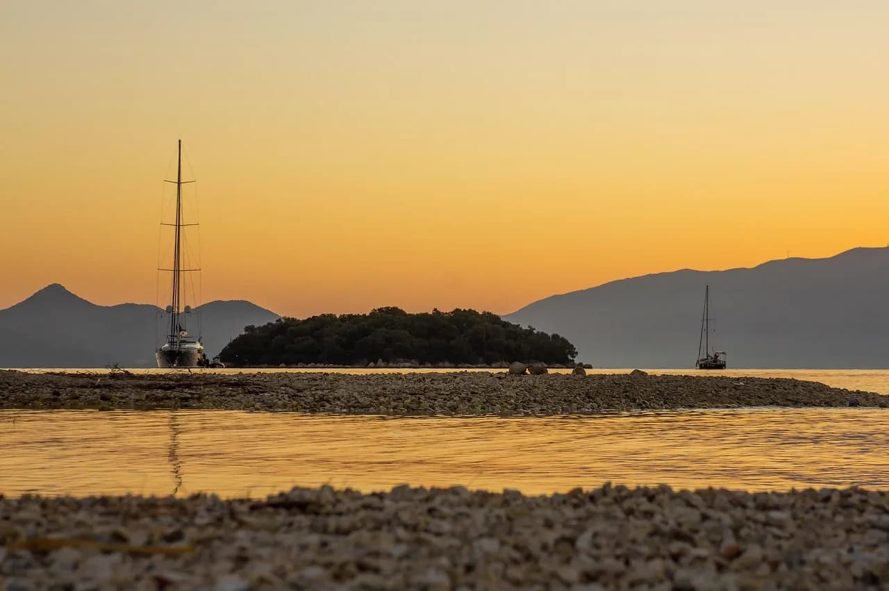 Two sailboats are silhouetted against a breathtaking sunset, with calm water and a small, tree-covered island in the distance.