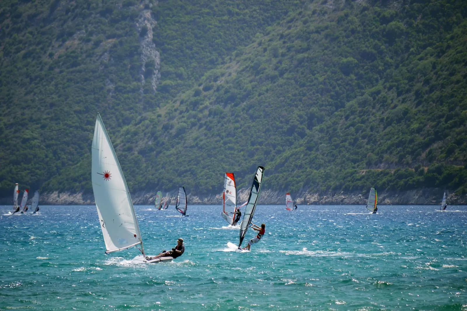 Windsurfers enjoy the windy conditions on the clear blue waters of Lefkada, set against a backdrop of a green, mountainous coastline.