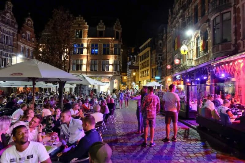 Locals and tourists fill the outdoor seating of pubs and restaurants along a vibrant and illuminated street in Leuven's city center at night.