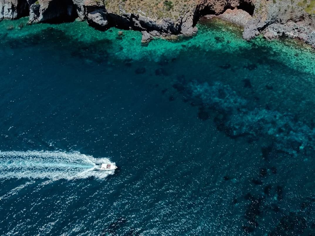 An aerial view of a boat cruising on the deep blue sea, with the rocky, green coastline in the background.