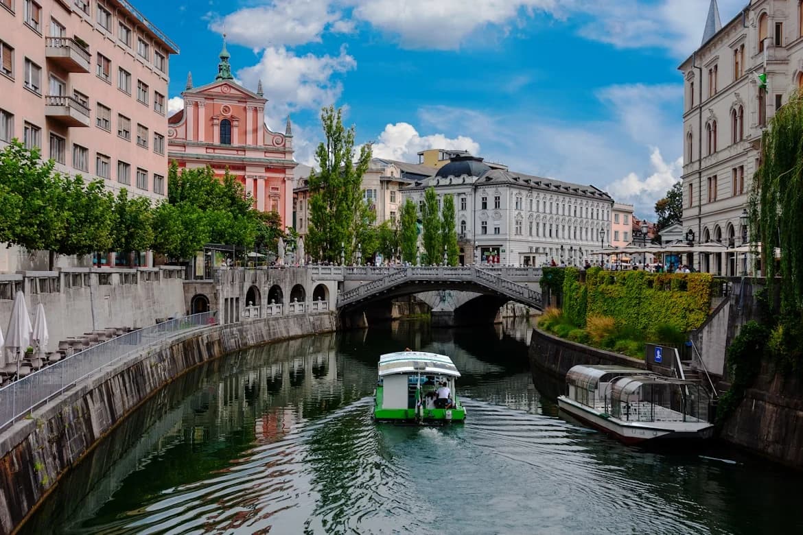 A small tourist boat cruises down the Ljubljanica River, with the pink facade of the Franciscan Church of the Annunciation and historic bridges in the background.
