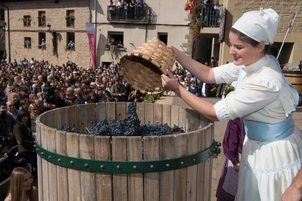 A woman in traditional attire empties a basket of grapes into a large wooden barrel, a local festival tradition.
