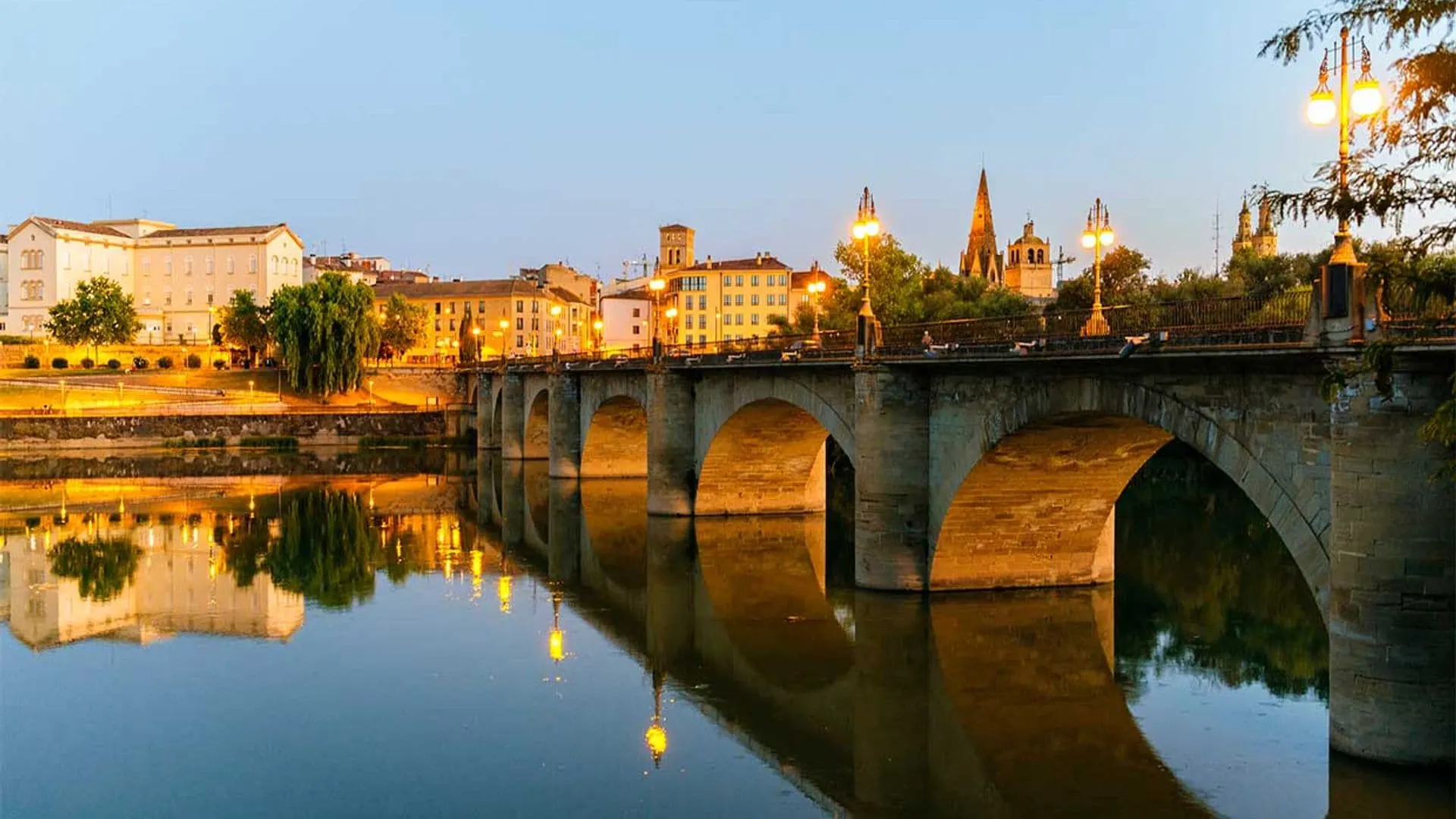 The Puente de Piedra, a historic stone bridge with illuminated streetlights, crosses the Ebro River at dusk.