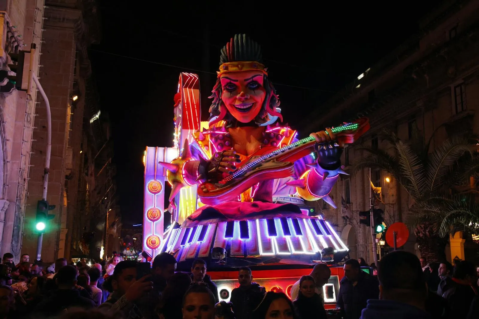 A large, elaborate float of a cartoonish queen playing a keyboard is a highlight of a festive carnival procession at night.