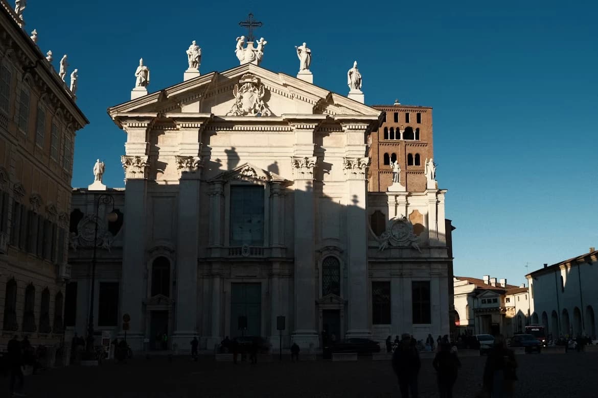 The sun casts a long shadow over the elegant facade of the Basilica of Sant'Andrea, with its statue-topped roof and a clock tower in the background.
