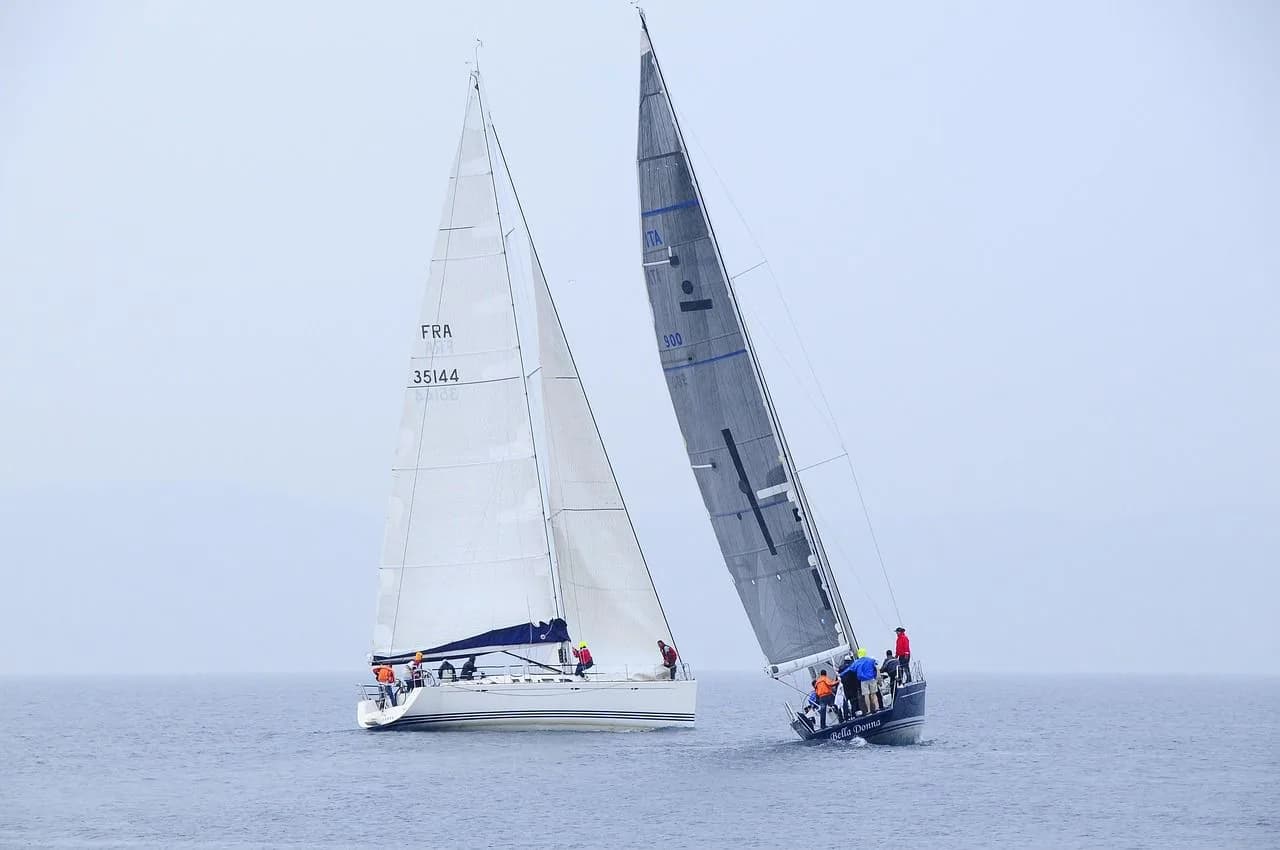 Two sailboats race on the open sea under a cloudy, white sky.