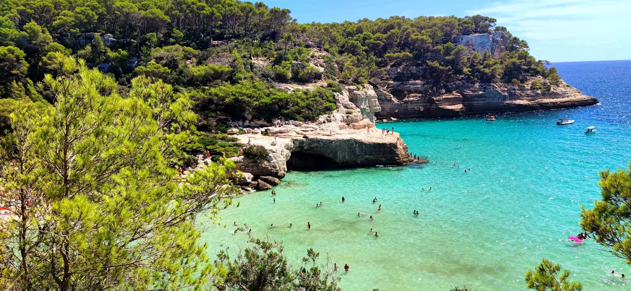 A hidden beach with crystal-clear turquoise water is nestled between two rocky hillsides covered in lush green trees.