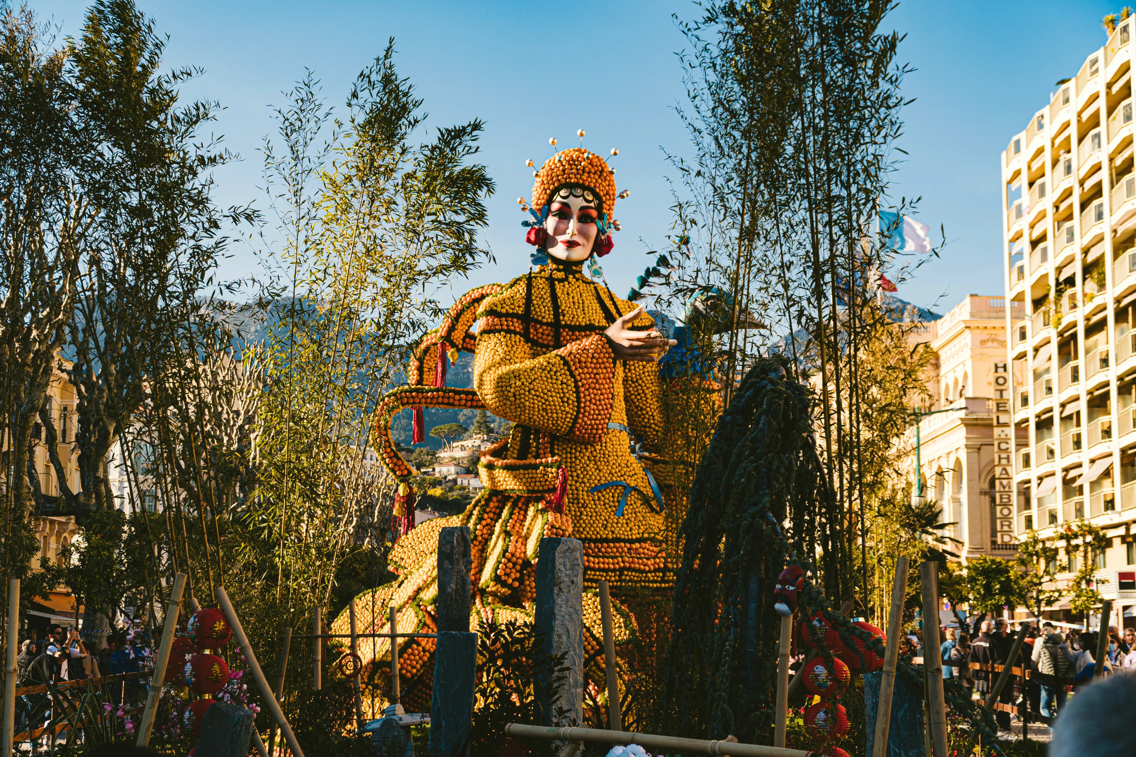 A large, ornate statue of a woman made of lemons and oranges is a centerpiece of a festival, with people and buildings in the background.