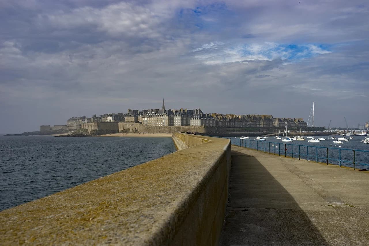A wide-angle view of a seaside promenade shows the rugged, rocky coastline and a historic city wall with the sea in the background.