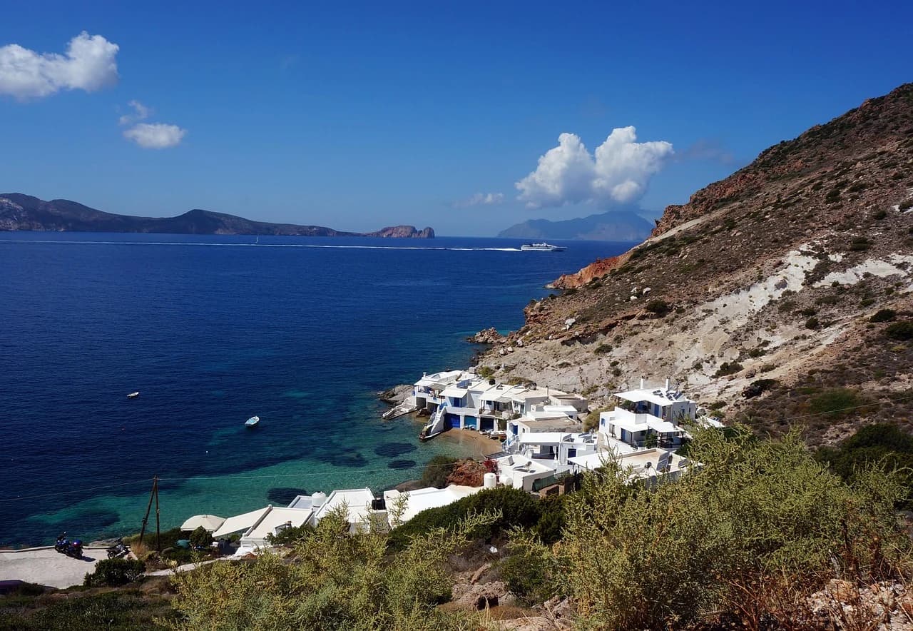 A tranquil bay is filled with clear blue water and surrounded by rocky cliffs, with a few white buildings and a cruise ship in the distance.