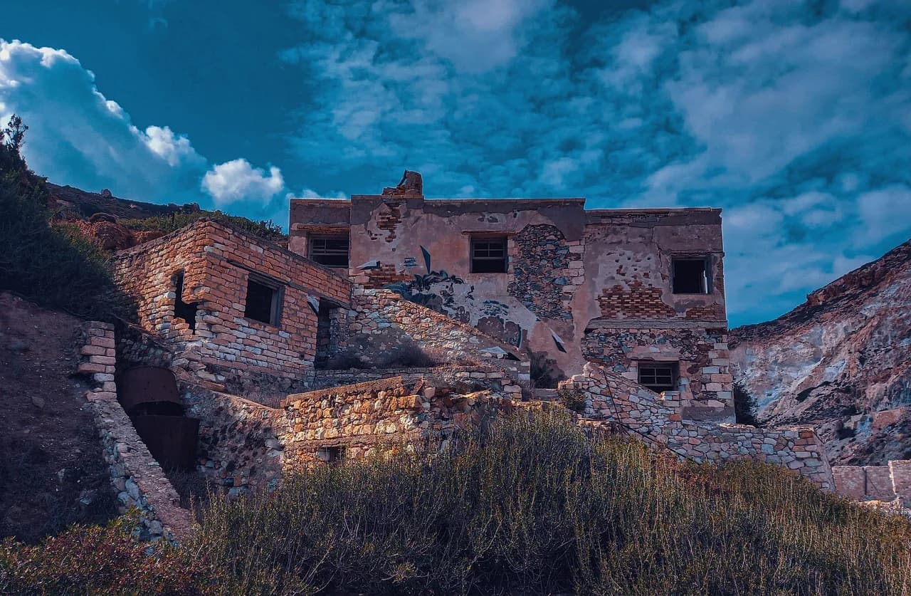 The weathered, rustic ruins of an old building stand on a rocky hillside, with a stunning blue sky and dramatic cliffs.