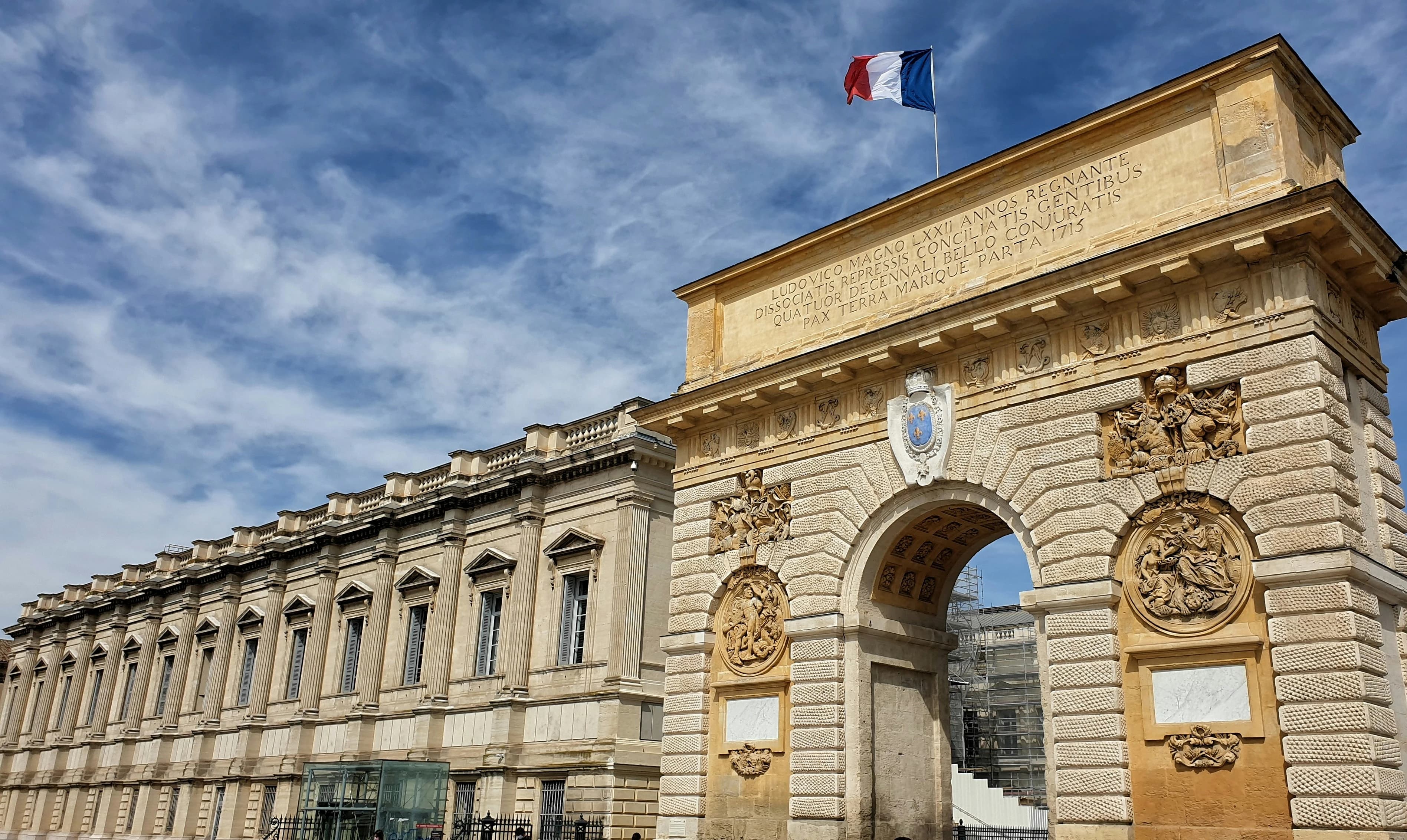 The Arc de Triomphe de Montpellier, a historic archway with intricate carvings, stands at the entrance to a city park.
