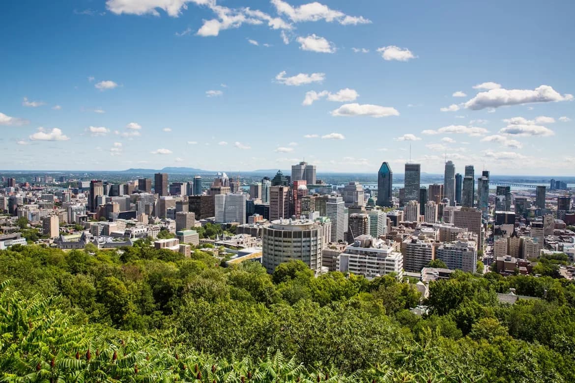An aerial view of the Montreal city skyline shows a dense cluster of modern skyscrapers and buildings, with a large, green park in the foreground.