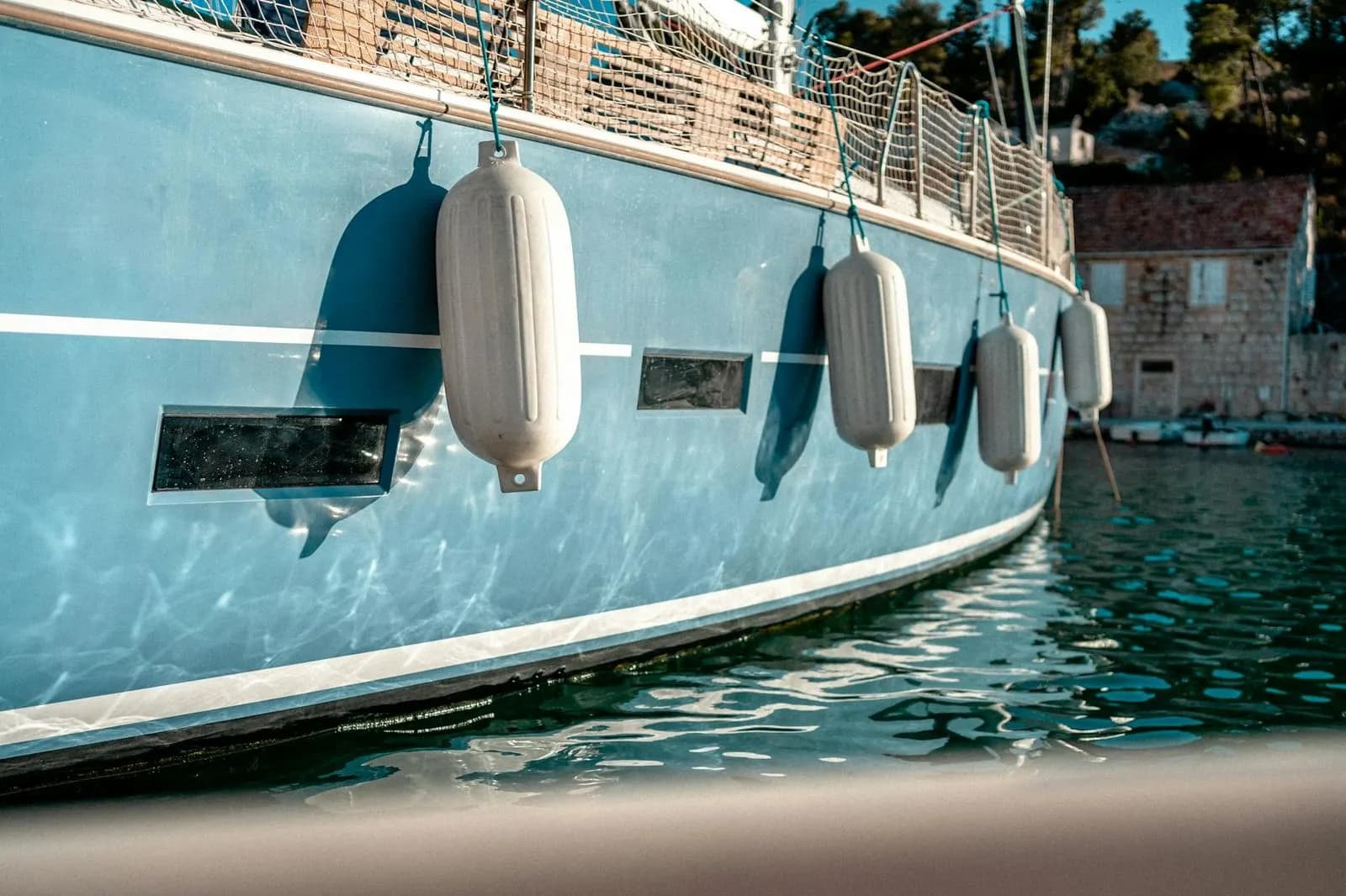 A close-up of a blue sailboat shows a series of white fenders hanging from the side, with a historic building on a hill in the background.