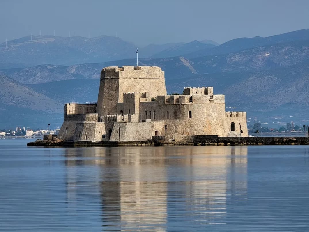 The historic Bourtzi Castle, a stone fortress on a small island, is viewed from a distance on the calm, blue sea.