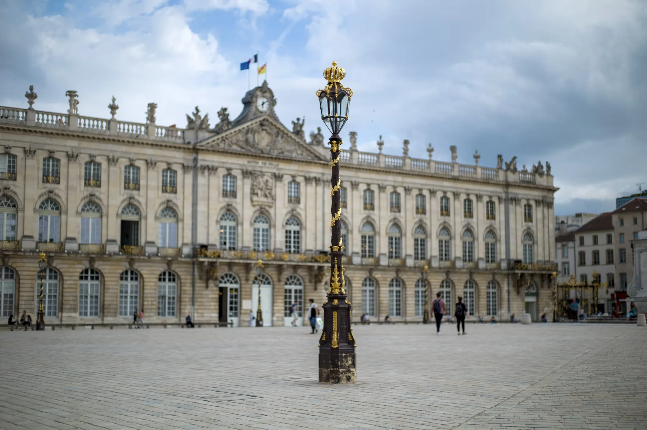 A golden, ornate lamppost stands in the foreground, with the grand facade of the City Hall of Nancy in the background.