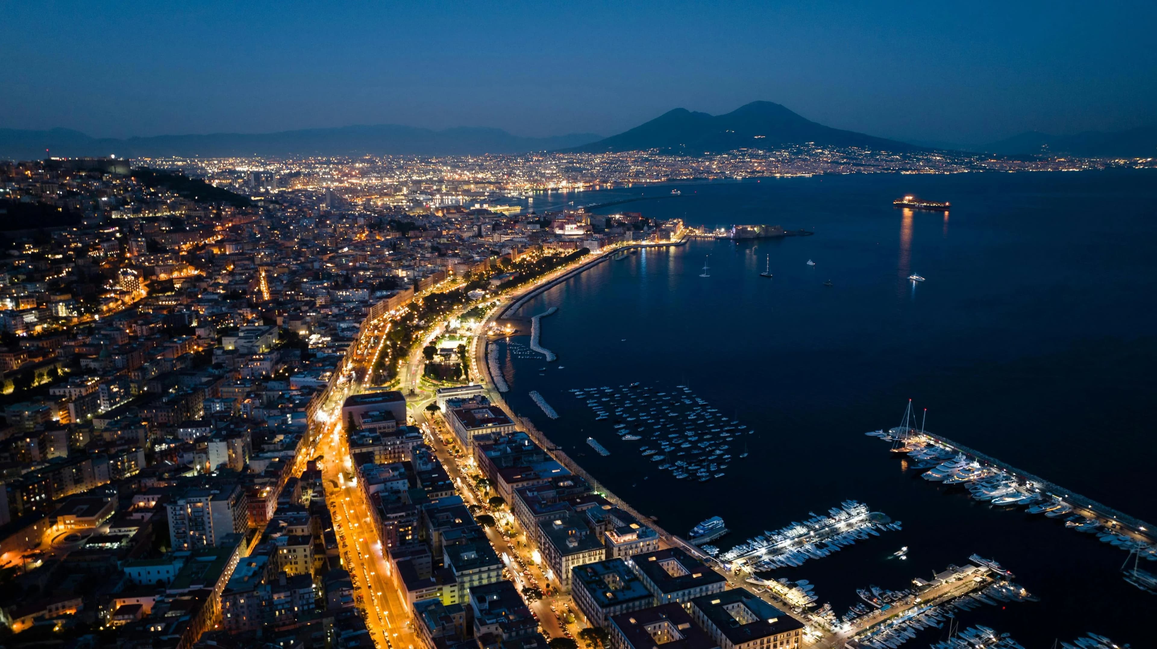 An aerial view of Naples at dusk shows the city lights and a wide bay with sailboats and a distant view of Mount Vesuvius.