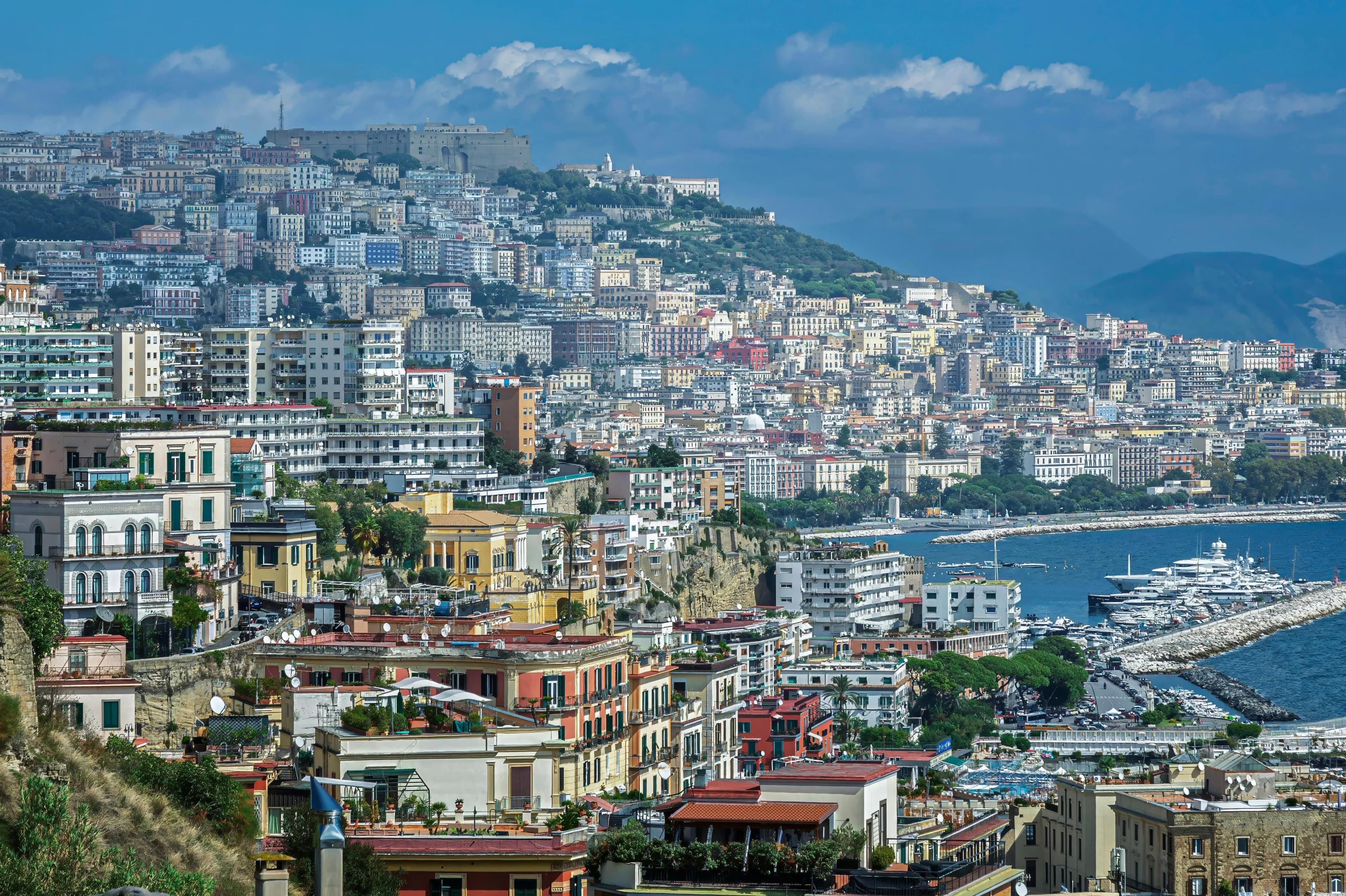 A panoramic view of Naples shows the dense residential buildings climbing up the hillside, with a busy harbor and the sea below.