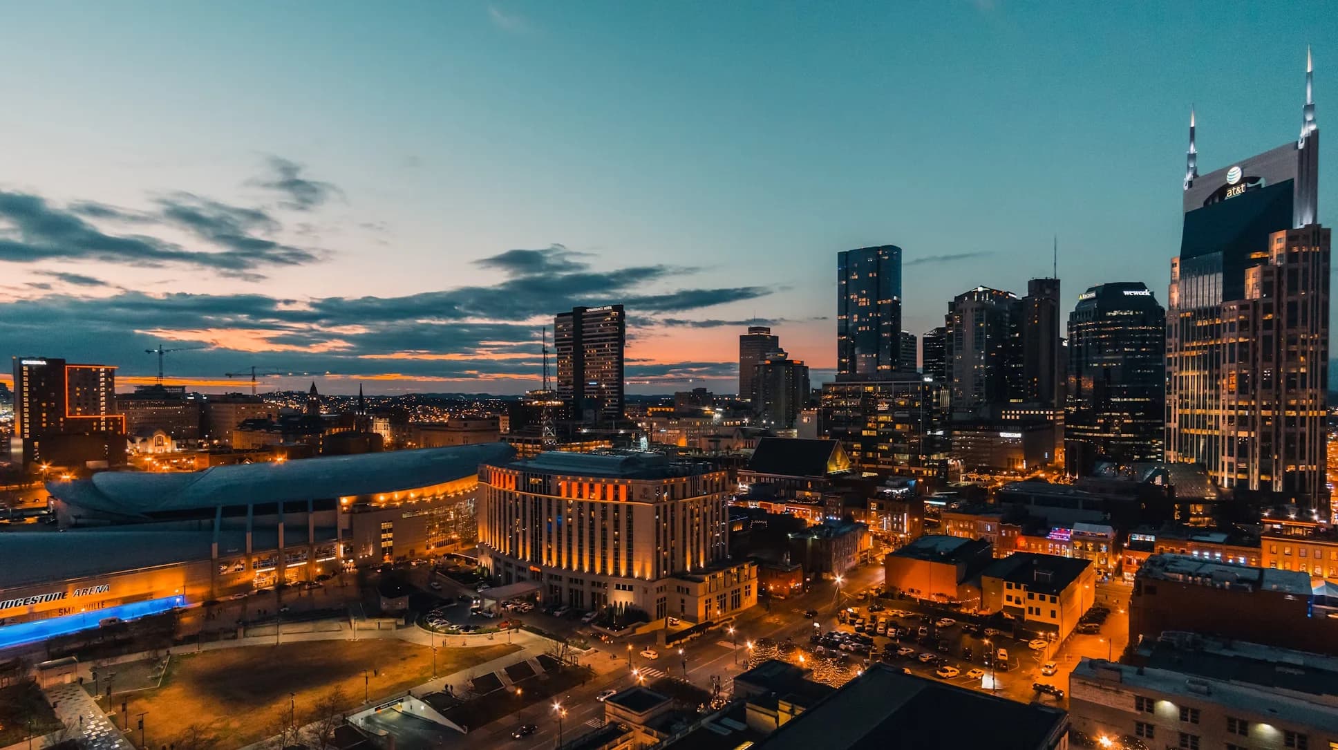 An aerial view of the Nashville skyline at dusk, with the city lights glowing and the Cumberland River flowing below.