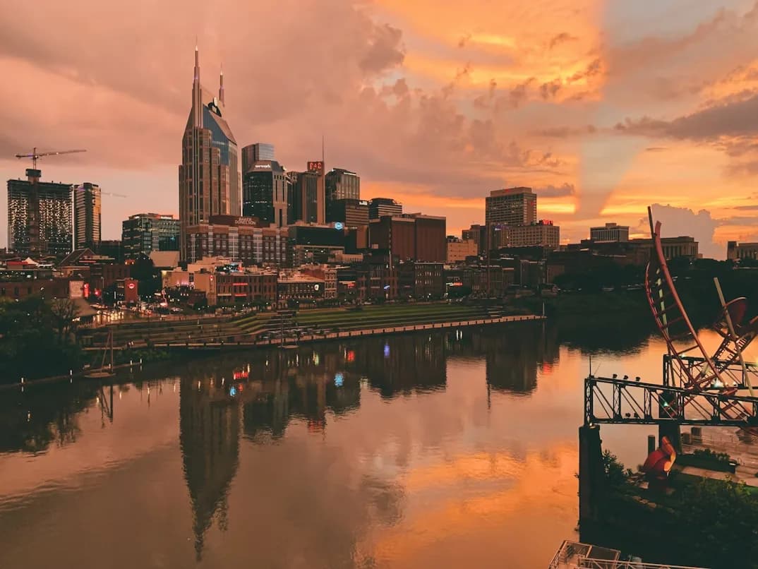 The skyline of Nashville is reflected on the Cumberland River at sunset, with the city lights and a unique sculpture in the foreground.