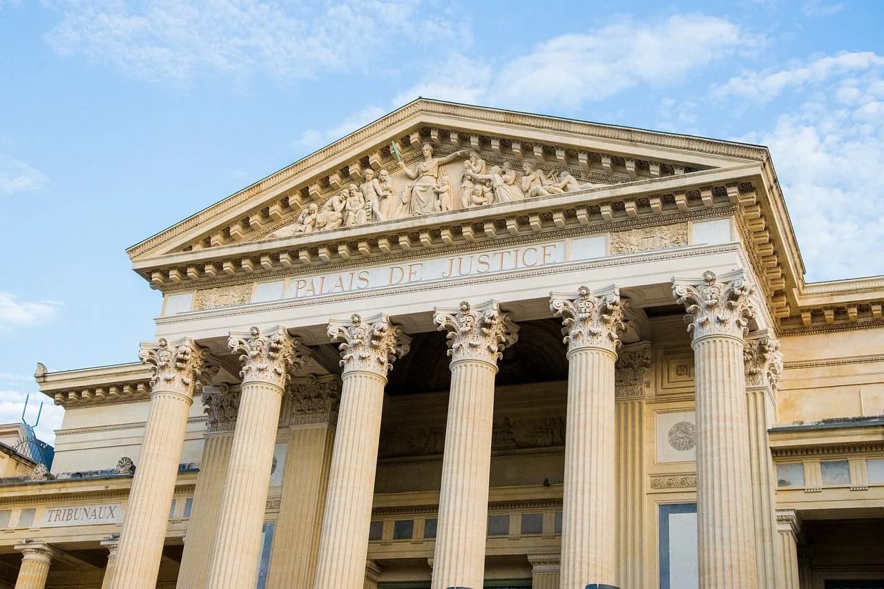 The grand, neoclassical facade of the Palais de Justice, a courthouse with large columns and a relief sculpture, stands against a blue sky.