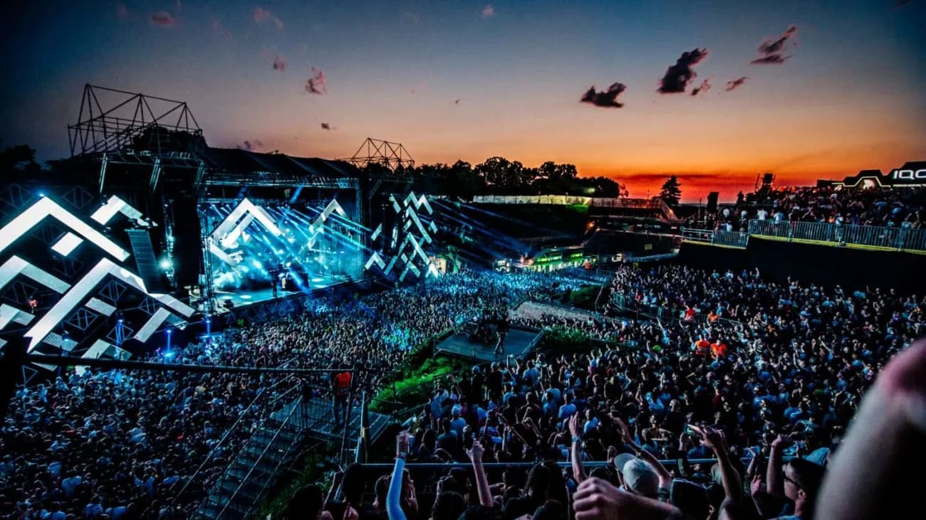 A music festival with a large stage and a vibrant crowd is set against the backdrop of a beautiful sunset.