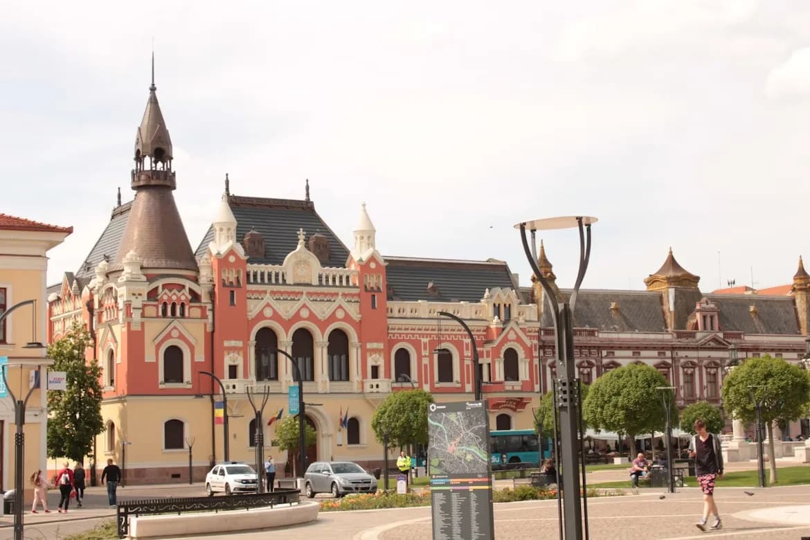 The ornate, pink and red facade of a historic building with a large steeple and numerous windows stands in a city square.